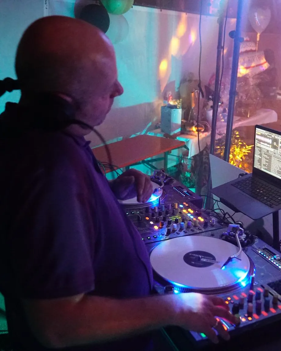 DJ an Plattenspielern und Mischpult, im Hintergrund Ballons, Geschenke und buntes Partylicht